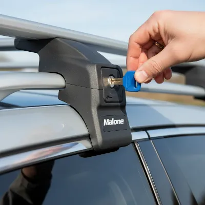 Close-up of a hand using a key to lock the integrated security mechanism on a Malone AirFlow2 crossbar foot, showing the clamp securely attached to a raised side rail, clear daylight, editorial style
